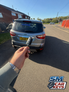 Replacement Ford EcoSport remote key held behind a grey Ford EcoSport after on-site key service by SM Car Keys.