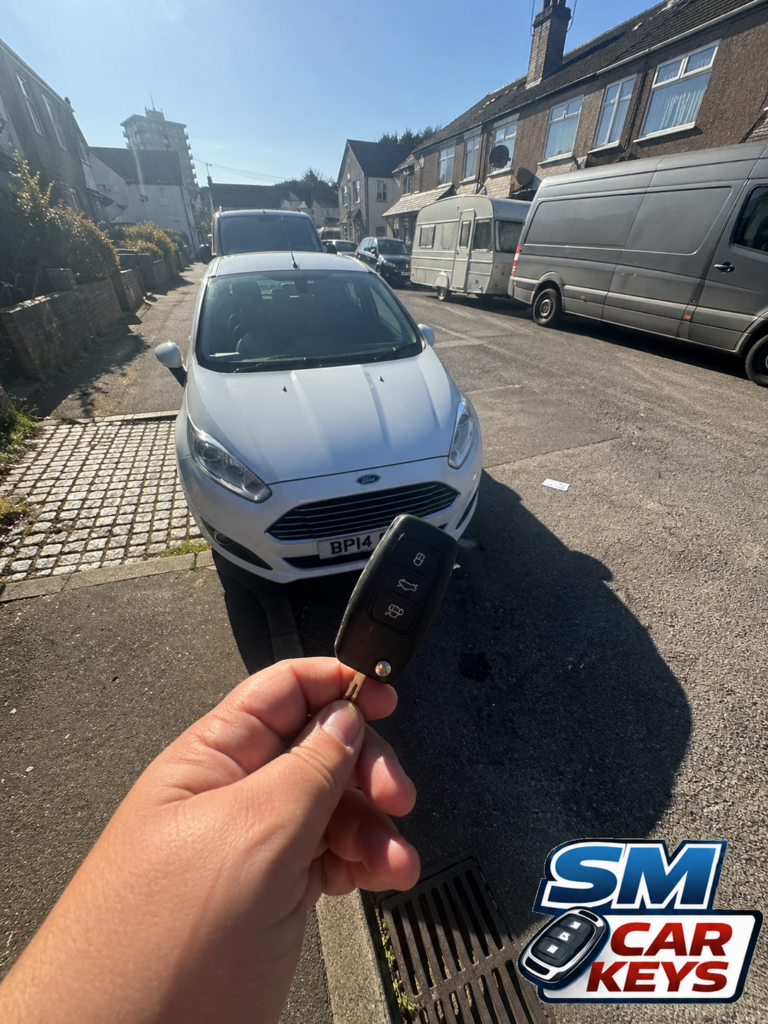 Replacement Ford Fiesta remote car key held in front of a white Ford Fiesta on a residential street by SM Car Keys mobile auto locksmith.